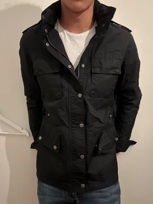 Svart field jacket med fickor - Snygg svart utilityjacka med flera stora fickor framtill och axelklaffar. Jackan har hög krage med tryckknappar och dragkedja, samt detaljer i metall. Perfekt för dig som gillar en stilren och funktionell look.