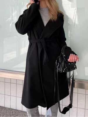 Missay Svart lång kappa med knytskärp - Stilren svart kappa i lång modell med bred krage och knytskärp i midjan. Kappan har en klassisk siluett och är tillverkad i ett mjukt tyg som ger en elegant look. Perfekt för kyligare dagar och enkel att matcha med olika outfits.