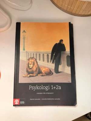 Psykologi 1+2a Lärobok - Läroboken 'Psykologi 1+2a' för gymnasiet av Martin Levander och Cornelia Sabelström Levander. Omslaget har en surrealistisk illustration med ett lejon och en person med svarta vingar. Boken har svart och orange färgtema och är i mjukt, matt material.