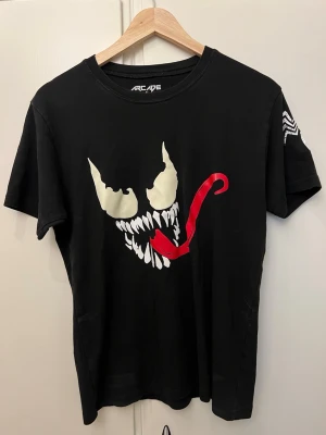 Svart Venom t-shirt från Arcade - Cool svart t-shirt från Arcade med ett stort Venom-tryck framtill. T-shirten är i 100% bomull och har en klassisk passform med korta ärmar. Perfekt för dig som gillar comics och vill sticka ut med en unik design.