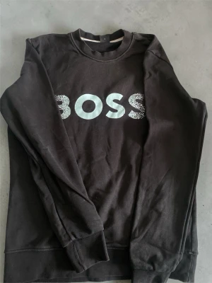 Svart sweatshirt med BOSS-tryck - Svart sweatshirt med rund hals och stort BOSS-tryck i vitt och ljusblått framtill. Tröjan har långa ärmar och ribbade muddar vid ärmslut och nederkant. Perfekt för en avslappnad och trendig look.