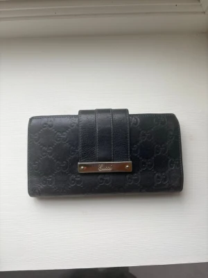 Svart plånbok från Gucci i läder - Snygg svart vintage 2011  plånbok från Gucci i äkta läder med präglat GG-mönster över hela ytan. Plånboken har en stilren design med ett brett band och en metallbricka framtill där Gucci-loggan syns. Perfekt för dig som gillar lyxiga accessoarer.