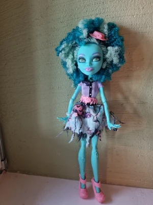 Monster High docka med blått hår - Säljer en Monster High docka med stort lockigt blått och vitt hår, ljusblå hud och detaljerade ansiktsdrag. Dockan har en lila topp, trasig kjol i svart, vitt och rosa samt rosa skor och en rosa liten hatt. Perfekt för samlare eller som present.