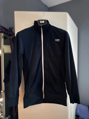 Mörkblå zip hoodie CORE by J&J - Snygg mörkblå hoodie från CORE by J&J med hel dragkedja framtill och hög krage. Tröjan har vita detaljer, långa ärmar och fickor på sidorna. Perfekt för en sportig och avslappnad stil.