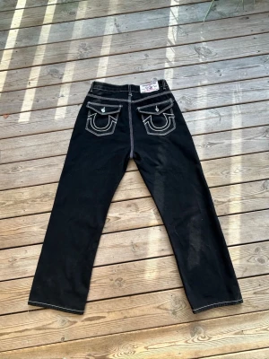 Feta true religion jeans svart o vita - Säljer ett par svarta jeans med raka ben och tydliga vita kontrastsömmar. Baksidan har två stora fickor med lock och dekorativa broderier. Jeansen har en klassisk passform och cool streetstil.  Jag är osäker på storlek men den är 38cm i midja 