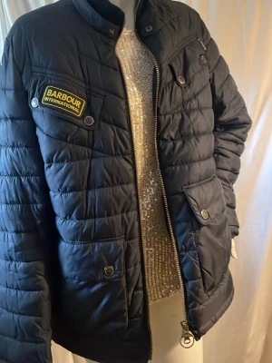 Svart pufferjacka från Barbour - Snygg svart pufferjacka från Barbour International med quiltad design och ståkrage. Jackan har flera fickor med tryckknappar, dragkedja framtill och en tydlig Barbour-patch   Obs finns väldigt liten fläck ej hål syns knappt vid typ handled se bild !! Annars mkt bra skick original påse finns till den se bild 