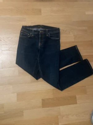 Mörkblå nudie jeans med gul söm - 29/32 Säljer ett par mörkblå jeans med klassisk femficksmodell och snygga guldfärgade sömmar på bakfickorna. Jeansen har normal midja och raka ben. Materialet är denim och de har en tidlös look som passar till det mesta.