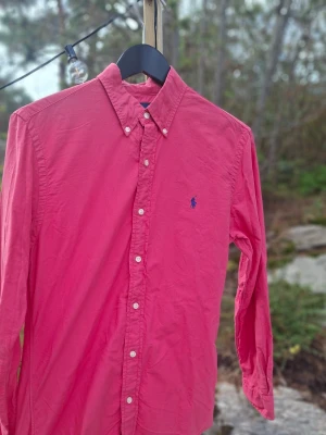 Rosa skjorta med blå logga - Säljer en klassisk rosa skjorta med button down-krage och blå broderad logga på bröstet. Skjortan har långa ärmar och vita knappar framtill. Perfekt för dig som gillar stilrena plagg med en färgklick.