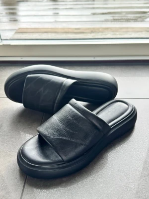 Svarta slip-in sandaler i skinnimitation - Stilrena svarta slip-in sandaler med bred rem och chunky sula. Tillverkade i skinnimitation med mjuk innersula för extra komfort. Perfekta för en avslappnad look och passar till det mesta i garderoben.
