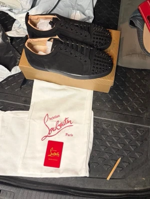 Svarta Louboutin sneakers med nitar - Säljer ett par svarta sneakers från Christian Louboutin med mattsvarta nitar på tån och sidorna. Skorna har en låg profil, svarta skosnören och en klassisk röd detalj undertill. Kommer med originalpåse och box.