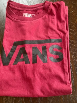 Röd Vans t-shirt med logga - Snygg röd t-shirt från Vans med stor svart logga framtill. Klassisk rund halsringning och korta ärmar. Tillverkad i mjuk bomull som känns skön mot huden. Perfekt för dig som gillar streetwear och vill ha en enkel men stilren look. Storlek L i barn