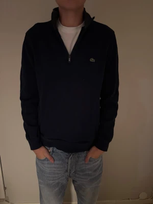 Mörkblå half-zip tröja från Lacoste - Mörkblå långärmad half-zip från Lacoste med hög krage och dragkedja framtill i storlek L. Tröjan är använd ett fåtal gånger och har en fungerande dragkedja med en liten broderad krokodil-logga på bröstet och ribbade muddar vid ärmslut och nederkant. Perfekt för lager på lager och en clean look.