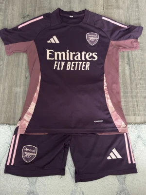 Lila Arsenal träningsset från Adidas - Säljer ett lila Arsenal träningsset från Adidas med tröja och shorts. Setet har ljusrosa detaljer och tryck med 'Emirates Fly Better' på tröjan. Perfekt för fotbollsträning eller sportiga dagar. Materialet är lätt och andas bra.