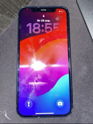 Iphone 12 - Säljer en mörkblå iPhone med stilren design och rundade hörn. Telefonen har en stor skärm med tunn ram och Face ID. Baksidan är i glas och har två kameror samt blixt i hörnet. Perfekt för dig som gillar Apple och vill ha en snygg smartphone.
