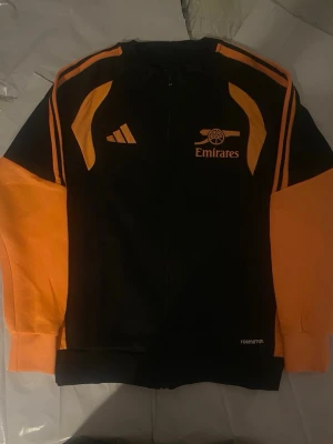 Svart och orange Arsenal träningsjacka - Snygg svart träningsjacka från Adidas med orange detaljer och Arsenal-logga samt Emirates-tryck på bröstet. Jackan har dragkedja framtill och är tillverkad i ett sportigt material, perfekt för träning eller fotbollsentusiasten.