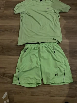 Grönt träningsset från Under Armour - Snyggt grönt träningsset från Under Armour med t-shirt och shorts. Setet är gjort i ett lätt och snabbtorkande material, perfekt för träning. T-shirten har rund hals och logga på bröstet, shortsen har resår i midjan, dragkedjefickor och logga på benet.