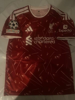Liverpool röd matchtröja Adidas - Snygg röd Liverpool matchtröja från Adidas med vita detaljer och tryck. Tröjan har klubbmärke, sponsorloggor och Champions League-märke på ärmen. Tillverkad i lätt och ventilerande material, perfekt för fotboll eller träning.