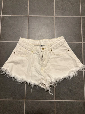 Vita fransiga shorts från H&M Divided - Säljer ett par vita shorts från H&M Divided med fransig kant nertill och klassisk femficksmodell. De har hög midja och stängs med dragkedja och knapp. Perfekta för sommaren och lätta att matcha med olika toppar.