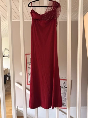 Vinröd långklänning med meshdetalj - Säljer en elegant vinröd långklänning med en axelbandslös design och meshdetalj med små pärlor på ena axeln. Klänningen har en oneshoulder-look och faller snyggt längs kroppen. Perfekt för bal eller festliga tillfällen. Klänningen är stretch och passar även för personer med storleken L