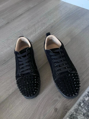  Louboutin svarta  - Säljer ett par svarta sneakers i mocka med svarta nitar på tån och svarta skosnören. Skorna har en låg profil och en stilren design med coola detaljer som sticker ut. Perfekta för dig som vill ha något unikt och edgy till din outfit.