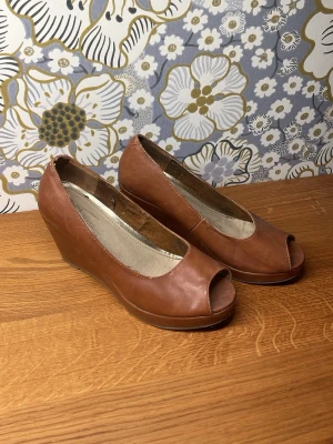 Bruna kilklackade peep toe pumps - Snygga bruna pumps med kilklack och öppen tå. Skorna har en klassisk design med rundad öppning framtill och är tillverkade i ett slätt, läderliknande material. Perfekta för dig som vill ha både höjd och komfort i din stil.