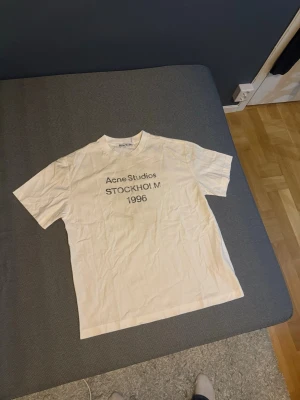 Vit Acne Studios t-shirt Stockholm - Vit t-shirt från Acne Studios med tryck 'Acne Studios STOCKHOLM 1996' på bröstet. Klassisk rak passform och rund halsringning. Perfekt för dig som gillar minimalistisk och stilren design.