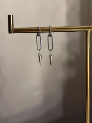 Silvriga örhängen med spike-detalj - Säljer ett par stilrena örhängen i silverfärg med avlånga länkar och spetsiga spike-hängen. De har en modern och edgy look som passar perfekt till dig som gillar minimalistiska accessoarer med attityd.