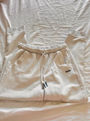 Vita mjukisbyxor från BOSS - Säljer ett par vita mjukisbyxor från BOSS med dragsko i midjan och två sidofickor. Byxorna har ribbade muddar vid bensluten och en clean look med BOSS-logga broderad på ena benet. Perfekta för chill dagar eller när du vill vara bekväm.