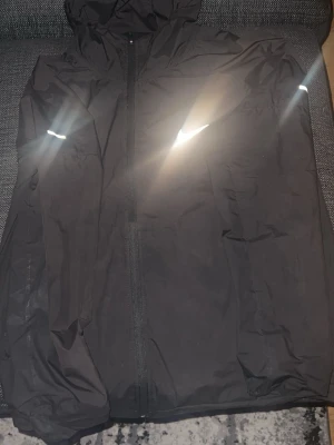 Svart Nike miller - Snygg svart Nike miller med huva och hel dragkedja framtill. Jackan har reflekterande detaljer på ärmarna och bröstet samt en minimalistisk design. Perfekt för blåsiga dagar och enkel att matcha med olika outfits.