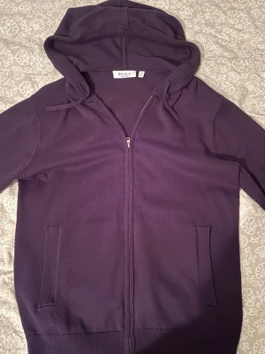 Mörklila hoodie med dragkedja RYVLS - Mörklila hoodie från RYVLS i storlek S. Tröjan har dragkedja framtill, två fickor och justerbar huva med snören. Tillverkad i mjukt material som passar perfekt för chill dagar. Enkel och stilren design utan tryck eller mönster.