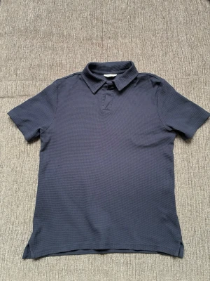 Mörkblå pikétröja East West - Mörkblå pikétröja från East West i strukturerad bomull. Klassisk krage, korta ärmar och enkel design utan tryck. Perfekt för en stilren och avslappnad look.