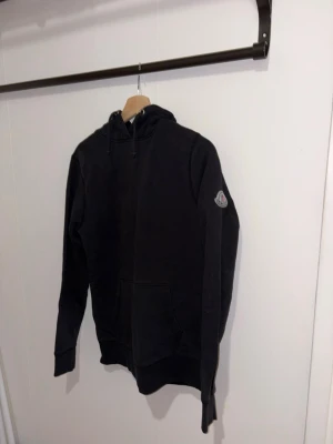 Svart moncler hoodie - Svart hoodie från Moncler med dragkedja framtill, huva med dragsko och klassisk logga på ärmen. Tillverkad i mjukt material som känns skönt mot huden. Perfekt för en chill och stilren look.