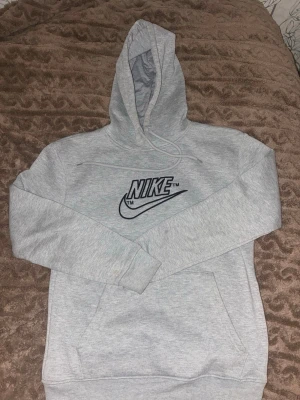 Grå hoodie från Nike med logga - Snygg grå hoodie från Nike med stor svart broderad logga på bröstet. Klassisk känguruficka framtill och justerbar huva med dragsko. Tillverkad i mjukt sweatshirtmaterial som är perfekt för chill eller streetwear.