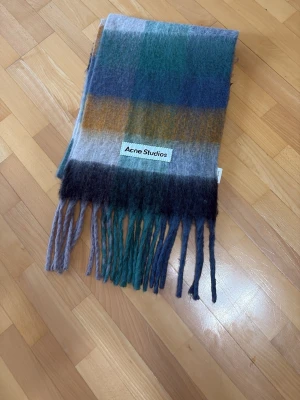 Rutig halsduk från Acne Studios - Säljer en snygg rutig halsduk från Acne Studios i mjuk ullblandning. Färgerna går i blått, grönt, grått och brunt med breda blockränder och långa fransar i ändarna. Perfekt accessoar för att lyfta din outfit.