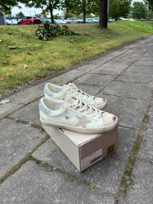 Feta Beige golden goose för damer - Köpte till syrran för 4200 men hon gillar inte dem. Säljer ett par vita Golden Goose sneakers med beige stjärna på sidan och glitterdetaljer på hälen. Skorna har beige snörning och slitna detaljer för en trendig look. Tillverkade i läder och textil, levereras med originalkartong och dustbag.