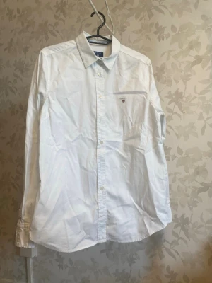 Gant Vit klassisk skjorta med knappar - Säljer en Gant  vit skjorta med klassisk krage och knäppning framtill. Skjortan har lång ärm och en liten broderad logga på bröstet. Perfekt till jeans eller chinos för en clean look.