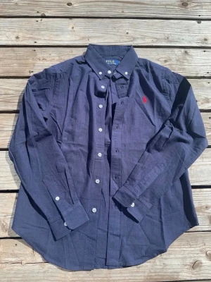 Mörkblå skjorta från Polo Ralph Lauren - Klassisk mörkblå skjorta från Polo Ralph Lauren med röd broderad logga på bröstet. Skjortan har lång ärm, knappar framtill och button-down krage. Tillverkad i mjuk bomull som känns skön mot huden. Perfekt för en clean och stilren look.