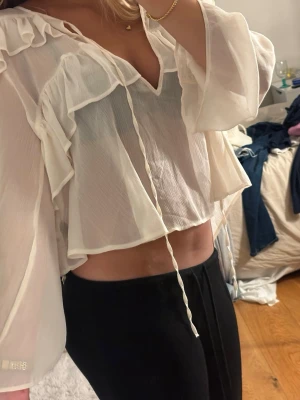 Vit volangblus med långa ärmar - Snygg vit blus i transparent, lätt material med volangdetaljer och knytband framtill. Blusen har v-ringning och långa, vida ärmar som ger en romantisk och luftig känsla. Perfekt att styla med jeans eller kjol för en trendig look. Aldrig använd ❤️