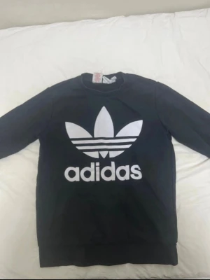 Svart sweatshirt från Adidas - Svart sweatshirt med stor vit Adidas Trefoil-logga och text på bröstet. Klassisk rund halsringning och långa ärmar. Tillverkad i mjukt material som passar perfekt till en avslappnad streetwear-look.