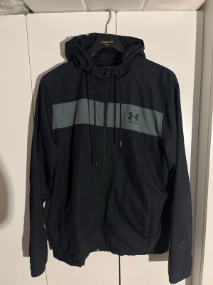 Svart vindjacka från Under Armour - Svart vindjacka från Under Armour med grått horisontellt band över bröstet och logga på vänster sida. Jackan har huva med dragsko, hel dragkedja och två sidofickor. Lätt material, perfekt för blåsiga dagar.
