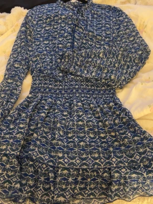Blåvit mönstrad klänning från Zara - Säljer en blåvit kortklänning från Zara med lång ärm och smock i midjan. Klänningen har ett grafiskt mönster, volangdetalj och knyt framtill. Tillverkad i ett lätt och luftigt material, perfekt för vår och sommar.