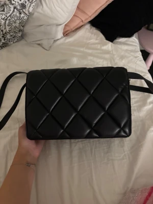 Svart axelväska - Snygg svart axelväska i quiltat mönster med rektangulär form och bred axelrem. Väskan är gjord i ett glansigt material som ser ut som konstläder. Perfekt för dig som gillar stilrena accessoarer med trendig touch. Jätte fin!!❣️