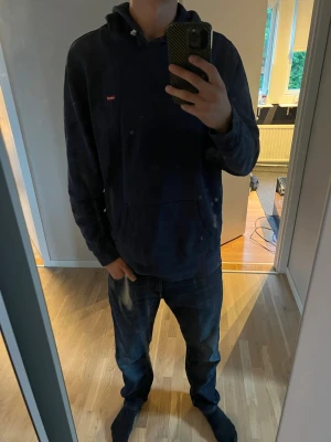 Mörkblå hoodie med liten logga - Säljer en mörkblå hoodie med huva och magficka. Tröjan har en liten röd logga på bröstet och dragsko i huvan. Perfekt för dig som gillar en enkel och stilren look. Tillverkad i mjukt material som känns skönt mot huden.