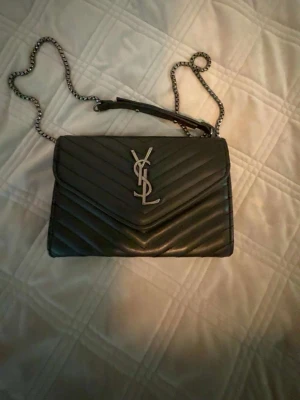 Svart YSL axelväska i läder - Säljer en svart axelväska från YSL i quiltat läder med silverfärgad kedjerem och ikonisk YSL-logga framtill. Väskan har ett stilrent och klassiskt utseende med v-format mönster på framsidan. Perfekt för dig som vill ha en lyxig accessoar.