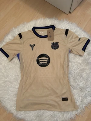 Beige FC Barcelona matchtröja Nike - Snygg beige FC Barcelona matchtröja från Nike med svarta detaljer och klubbmärke på bröstet. Tröjan har korta ärmar, rund hals med blå och svarta inslag samt tryck från Spotify och UNHCR. Tillverkad i lätt och ventilerande material, perfekt för fotboll eller träning.