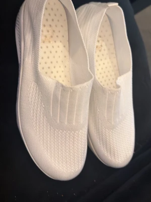Vita slip-on sneakers i mesh - Fräscha vita slip-on sneakers med ovandel i ventilerande mesh och elastisk resår över vristen. Sulan är vit med en synlig luftbubbla på sidan för extra cool känsla. Perfekt för dig som gillar en clean och sportig stil.