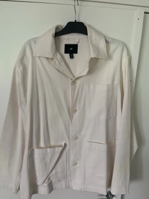 Vit skjortjacka från H&M - Stilren vit skjortjacka från H&M med avslappnad passform. Jackan har tre stora fickor framtill, knäppning med fyra knappar och klassisk krage. Tillverkad i ett lätt, vävt material som passar perfekt för lager på lager.
