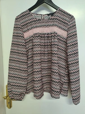Mönstrad blus med zigzag-mönster - en snygg blus med rosa och svart zigzag-mönster. Blusen har rund halsringning, långärmade ärmar med resår vid handlederna och en lös, flowy passform. Materialet känns lätt och luftigt, perfekt för en trendig vardagslook.rosa blå grå färger