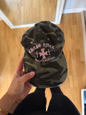 Camo keps med rosa brodyr - Säljer en camouflagemönstrad keps med rosa brodyr framtill där det står 'CHASE AFTER' och motiv av kors. Kepsen har böjd skärm och justerbar rem bak. Materialet är bomullstyg och modellen är klassisk dad cap.
