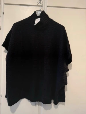 Svart stickad poncho med polokrage - En svart stickad poncho med hög polokrage och korta ärmar. Ponchon har en lös och avslappnad passform samt knappar längs sidorna. Perfekt för lager på lager och ger en stilren look till höst och vinter.